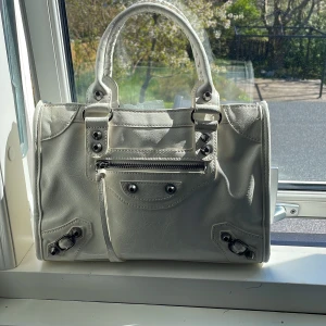 Vit handväska med silverdetaljer - Säljer en stilren vit handväska med silverfärgade metalldetaljer och dragkedja framtill. Väskan har två handtag och dekorativa spännen i hörnen. Perfekt för dig som vill ha en klassisk accessoar med coola detaljer.