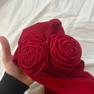 Röd långklänning med rosdetaljer - Säljer en röd långklänning med unika rosdetaljer vid bysten och snygg drapering längs sidan. Klänningen har halterneck och är axelbandslös, perfekt för dig som vill sticka ut på festen. Materialet känns mjukt och lyxigt.