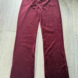 Vinröda mjukisbyxor från Gina Tricot - Mysiga vinröda mjukisbyxor från Gina Tricot med dragsko i midjan. Byxorna har raka ben och är tillverkade i ett mjukt, sammetsliknande material. Perfekta för en avslappnad stil.