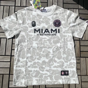 Vit camo t-shirt från A Bathing Ape x Inter Miami - Säljer en vit och ljusgrå camo t-shirt från A Bathing Ape i samarbete med Inter Miami. T-shirten har tryck med MIAMI och BAPE-logga fram, samt stort BAPE 93-tryck bak. Rund hals och kort ärm. Perfekt för dig som gillar streetwear och fotboll. Perfect för sommaren 