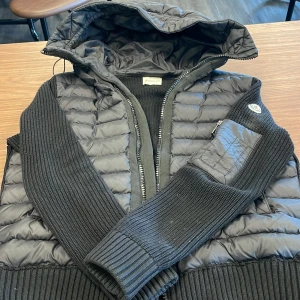 Svart dunväst med stickade ärmar från Moncler - Snygg svart dunväst från Moncler med stickade ärmar och huva. Västen har dragkedja framtill och en liten ficka med dragkedja på ena ärmen. Perfekt för lager-på-lager och stilren look.