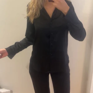 Svart skjorta med knappar - Stilren svart skjorta med klassisk krage och knäppning framtill. Skjortan har långa ärmar och en rak passform, perfekt för en clean och enkel look. Passar till många olika outfits och är lätt att matcha.