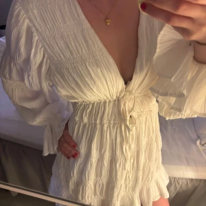 Vit omlottklänning med volang från NA-KD - Superfin vit kortklänning från NA-KD med omlottmodell, volangdetaljer och knyt i midjan. Klänningen har långa, luftiga ärmar och en djup v-ringning. Perfekt för sommarens alla tillfällen! Kom gärna med prisförslag