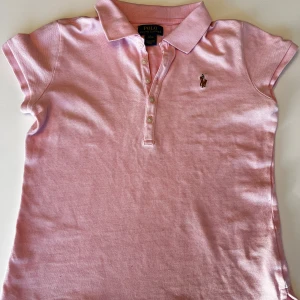 Ljusrosa pikétröja från Polo Ralph Lauren - Säljer en klassisk ljusrosa pikétröja från Polo Ralph Lauren med korta ärmar och knappslå framtill. Tröjan har broderad logga på bröstet och är tillverkad i bomull. Perfekt för en stilren och sportig look.