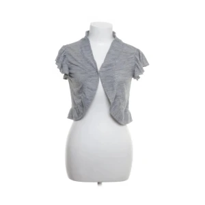 Grå bolerotopp från Pieces - Säljer en ljusgrå bolerotopp från Pieces med korta volangärmar och liten krage. Toppen är öppen framtill och har en mjuk, stickad känsla. Perfekt att slänga över en klänning eller topp för en extra touch.