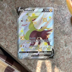 Pokémonkort Boltund V - Säljer ett holografiskt Pokémonkort med Boltund V. Kortet har en färgglad och glansig design med Boltund i fokus, HP 200 och el-typ. Text och attacker är tydliga på framsidan. Baksidan har klassisk blå Pokémon-design med Pokéboll.