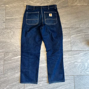 Blå jeansbyxor från Carhartt med raka ben - Säljer ett par klassiska blå jeansbyxor från Carhartt med raka ben och stora bakfickor. Byxorna har normal passform och är tillverkade i slitstarkt denim. Perfekta för dig som gillar streetwear och funktionella detaljer.