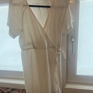 Vit omlottklänning från Vero Moda - Säljer en vit omlottklänning från Vero Moda med korta ärmar och knyt i midjan. Klänningen har en v-ringad omlottdesign och är tillverkad i ett lätt material, perfekt för varmare dagar.