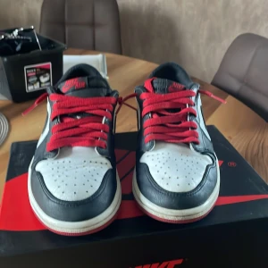 Nike Air Jordan 1 Low med röda snören - Säljer ett par Nike Air Jordan 1 Low med svart, vitt och rött läder samt röda snören. Klassisk vinge-logga på hälen och svart swoosh på sidan. Perfekta för dig som gillar streetwear och sneakers med ikonisk design.(logga där bak skrapad) 450 med låda!