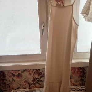 Vit långklänning med volang - Säljer en vit långklänning med smala axelband och volang nertill. Klänningen har en rak passform och är ärmlös, perfekt för varma dagar eller sommarens tillställningar.