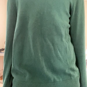 Grön stickad tröja - Mörkgrön stickad tröja med rund halsringning och långa ärmar. Tröjan har ribbade muddar vid ärmslut och nederkant, vilket ger en klassisk och enkel look. Perfekt för lager-på-lager under kyligare dagar.