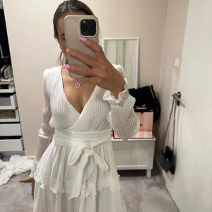 Vit omlottklänning med volang och knytband - Säljer en superfin vit omlottklänning med långa ärmar och volangdetaljer. Klänningen har ett knytband i midjan och v-ringad hals. Perfekt för sommarens tillfällen och studentfirande! Från Dennis maglic 