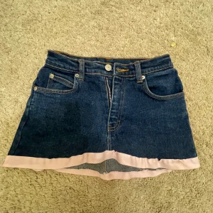 Jeanskjol med rosa kant från Cinema - Mörkblå jeanskjol från Cinema med fem fickor och dragkedja framtill. Kjolen har en ljusrosa kant längst ner som ger en unik touch. Klassisk denimlook med bälteshällor och bakfickor.