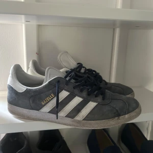 Adidas Gazelle grå/blå sneakers - Säljer ett par as feta Adidas Gazelle sneakers i grå mocka med vita detaljer och svarta snören. Skorna har de ikoniska tre ränderna på sidan och Adidas-logga på häl och plös. Perfekta för en stilren och sportig look. Säljer på grund av att de är för små. Storlek 44 men passar 43 bättre. Är öppen för byten i storlek 44-45