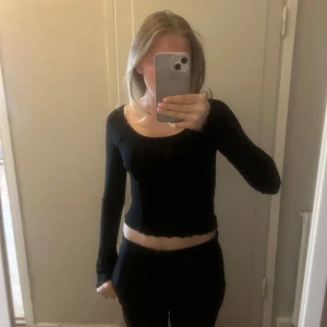 Svart  topp  - Säljer en svart långärmad croppad topp från Bershka. Toppen har en bred, rundad urringning och dekorativa sömmar framtill. Materialet är stretchigt och sitter tight mot kroppen. Perfekt till jeans eller kjol!