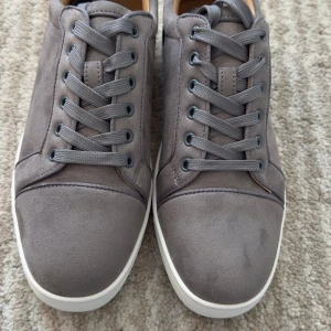 Gråa sneakers med röd sula - Säljer ett par stilrena gråa sneakers från Christian Louboutin med klassisk röd sula. Skorna har snörning, rund tå och är tillverkade i mocka med vita kanter runt sulan. Perfekta för dig som vill sticka ut med en lyxig detalj.