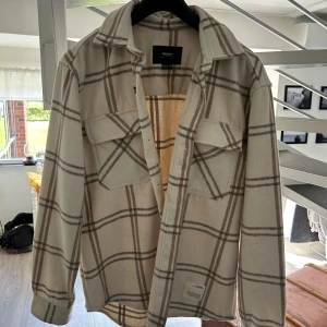Beige rutig overshirt från Pull&Bear - Säljer en beige overshirt från Pull&Bear med rutigt mönster i brunt och grått. Skjortan har två bröstfickor med lock och knappar framtill. Perfekt att bära som lager över en t-shirt.