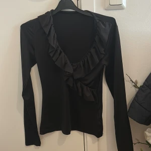 Svart långärmad topp med volang - Säljer en svart långärmad topp med djup v-ringning och volangdetaljer längs halsringningen. Snygg och stilren modell som passar perfekt till många olika outfits. Den är använd nån gång men annars helt ny.