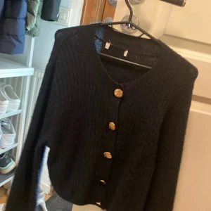 Svart stickad kofta med guldknappar - Säljer en svart stickad kofta med snygga guldknappar framtill. Koftan har lång ärm och ribbad struktur, perfekt att slänga på sig när det är kyligt. Enkel och stilren modell som passar till det mesta.