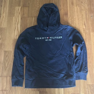 Mörkblå hoodie från Tommy Hilfiger - Säljer en mörkblå hoodie från Tommy Hilfiger med logga och texten 'EST. 1985' på bröstet. Tröjan har huva och långa ärmar, perfekt för en avslappnad stil. Klassisk och stilren design med mjukt material.
