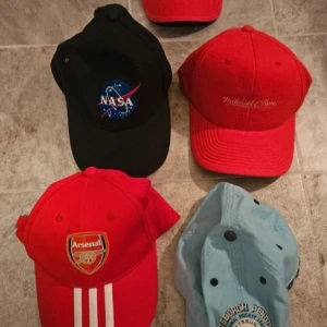 Flera kepsar med olika motiv och färger - Fem kepsar: en svart med NASA-logga, en röd med broderad text, en röd med Arsenal-märke och vita ränder, en ljusblå med broderad text och en enkel röd keps. Perfekt för dig som gillar sportiga och unika accessoarer.
