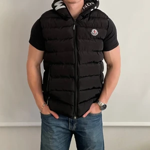 Svart dunväst från Moncler - Säljer en svart dunväst från Moncler med huva som inte är äkta. Den har en Moncler-logga på bröstet och en sportig, puffad design. Perfekt för lager-på-lager och snygg till jeans.