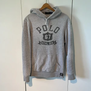 Grå Raffe hoodie - Säljer en klassisk grå hoodie från Polo Ralph Lauren med tryck 'POLO 67 ATHL. DIV.' på bröstet. Tröjan har huva och en stor magficka. Perfekt för en avslappnad stil.