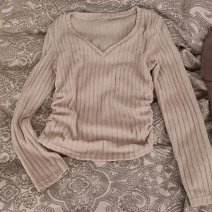 Beige ribbad långärmad topp från SHEIN - Säljer en beige ribbad långärmad topp från SHEIN i storlek M. Toppen har v-ringning och rynkade sidor som ger en snygg passform. Materialet är mjukt och stretchigt med inslag av viskos och polyester.