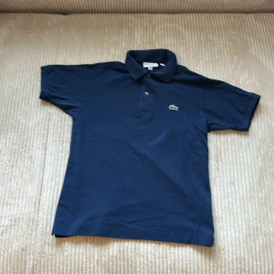 Mörkblå pikétröja från Lacoste - Säljer en klassisk mörkblå pikétröja från Lacoste med broderad logga på bröstet. Tröjan har korta ärmar, krage och knappar framtill.  100% äkta