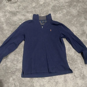Marinblå långärmad tröja från Ralph Lauren - Säljer en klassisk marinblå långärmad tröja från Ralph Lauren med halv dragkedja och liten broderad logga på bröstet. Perfekt för en stilren och avslappnad look.