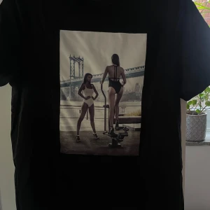Svart t-shirt med fotoprint från Limitato - Säljer åt min bror. Svart t-shirt från Limitato med ett stort fotoprint på framsidan som visar två kvinnor i badkläder framför en bro. T-shirten har klassisk passform och rund hals. Säljer med originallådan. Nypris ca 2700kr