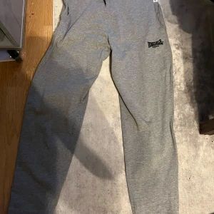 Grå mjukisbyxor från Lonsdale - Aldrig använda!!Snygga grå mjukisbyxor från Lonsdale med svart broderad logga på ena benet. Byxorna har en klassisk passform och är perfekta för chill eller träning. Mjukt material som känns skönt mot huden.