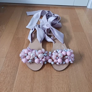 Tofflor från Elina Linardaki - Unika sandaler i ljuslila med breda sidenband att knyta runt ankeln. Framtill dekorerade med stora pärlor och glittrande stenar i rosa, lila och silver. Perfekta för dig som vill sticka ut med dina skor. Handgjorda av äkta läder i Grekland. Nypris ligger på 2500kr pris kan diskuteras.