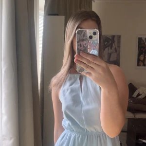 Ljusblå ärmlös klänning med nyckelhålsdetalj - Säljer en ljusblå ärmlös klänning med resår i midjan och en liten nyckelhålsöppning vid halsen. Klänningen har en mjuk, lätt glansig yta och faller fint. Perfekt för varma dagar eller speciella tillfällen.