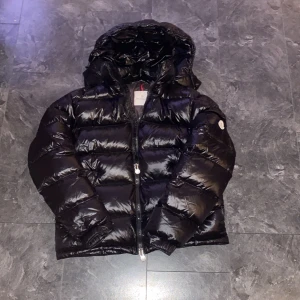 Moncler  - Säljer en svart dunjacka från Moncler med huva och dragkedja. Jackan har klassisk Moncler-logga på ärmen och detaljerade dragkedjor framtill. Fodret har ett serietidningsmönster på insidan. Perfekt för kyliga dagar.