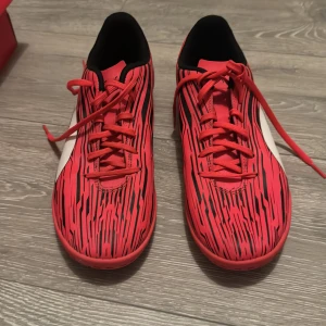 Röda fotbollsskor från Puma med svart mönster - Säljer ett par röda fotbollsskor från Puma med svart grafiskt mönster och vit logga på sidan. Skorna har snörning och rund tå, samt svart insida. Perfekta för dig som vill sticka ut på planen!