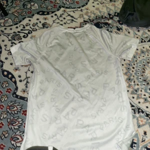 Vit PSG x Jordan t-shirt - Säljer en vit PSG t-shirt från Jordan med diskret Paris-tryck över hela tröjan och svart logga samt sponsortryck på bröstet. Röd PSG-logga på vänster sida. Perfekt för dig som gillar fotboll och streetwear.