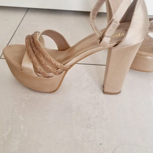 Beige platåsandaler med klack och glittriga band - Säljer ett par snygga beige sandaler med hög klack och platåsula. Skorna har glittriga band över foten och en öppen tå. Perfekta för dig som vill sticka ut lite extra!