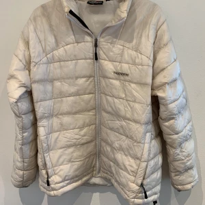 Vit quiltad jacka från TrueNorth - Säljer en vit quiltad jacka från TrueNorth med dragkedja framtill och hög krage. Jackan har en sportig look och är lätt vadderad, perfekt för kyligare dagar. Snygg och enkel design med diskret logga på bröstet. Finns hål i armhålen i insidan av jackan.