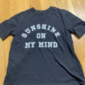 Svart t-shirt med texten 'Sunshine On My Mind' - Svart t-shirt med vit tryckt text 'SUNSHINE ON MY MIND' i stora bokstäver på framsidan. Klassisk rund hals och korta ärmar. Perfekt för en avslappnad stil.