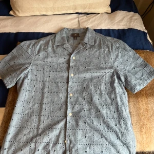 Grå kortärmad skjorta från H&M - Snygg grå skjorta från H&M med korta ärmar och ett unikt broderat mönster. Perfekt för en stilren look. Skjortan har knappar framtill och är tillverkad i ett lätt material som passar bra för varmare dagar. Har aldrig använt den 