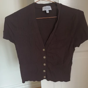 Brun kortärmad kofta från & Other Stories - Säljer en snygg brun kortärmad kofta från & Other Stories. Den har en v-ringad design med knappar framtill och är ribbad för en stilren look. Perfekt för att lägga till en touch av elegans till din outfit.
