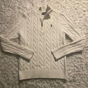 Vit stickad tröja från Ralph Lauren - Säljer en stilren vit stickad tröja från Ralph Lauren med kabelstickat mönster och en liten broderad logga på bröstet. Tröjan har en dragkedja vid halsen och ribbade muddar vid ärmslut och nederkant. Perfekt för kyliga dagar!
