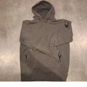 Grå hoodie med broderad detalj - Säljer en stilren grå hoodie med en liten broderad detalj på bröstet. Tröjan har en klassisk design med huva och fickor framtill. Perfekt för en avslappnad look.