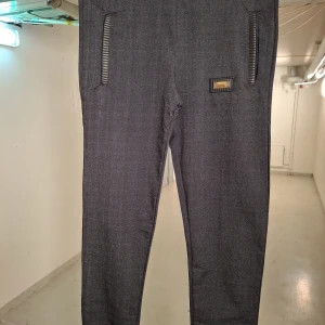 Mörkgrå byxor med dragkedjedetaljer - Snygga mörkgrå byxor med dragkedjor vid fickorna och en diskret logga på sidan. Byxorna har en modern och stilren design, perfekt för en avslappnad look. De är tillverkade i ett bekvämt material som ger en skön passform.
