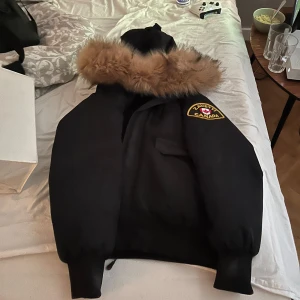 Svart vinterjacka från Zavetti Canada - Säljer en svart vinterjacka med pälskantad huva från Zavetti Canada. Jackan har ett quiltat foder och en broderad logotyp på ärmen. Perfekt för kalla dagar.