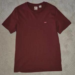 Vinröd t-shirt från Levi's - Säljer en snygg vinröd t-shirt från Levi's med en liten logga på bröstet. T-shirten är kortärmad och gjord i mjuk bomull. Perfekt för en avslappnad stil. Pyttelitet hål på mage som knappt syns. Hör av dig vid intresse!