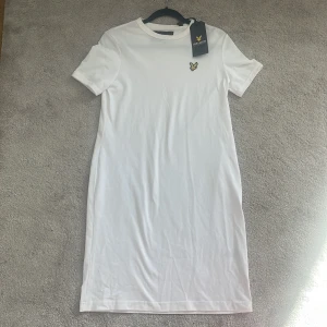 Vit klänning från Lyle & Scott - Snygg vit klänning från Lyle & Scott med korta ärmar och en broderad gul logga på bröstet. Perfekt för en stilren look. Klänningen har en enkel och klassisk design som passar till många tillfällen.