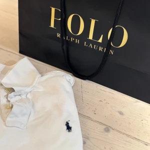 Vit pikétröja från Ralph Lauren - Säljer en klassisk vit pikétröja från Ralph Lauren med den ikoniska loggan broderad på bröstet. Tröjan har korta ärmar och en stilren krage med knappar. Perfekt för en avslappnad men ändå stilfull look.