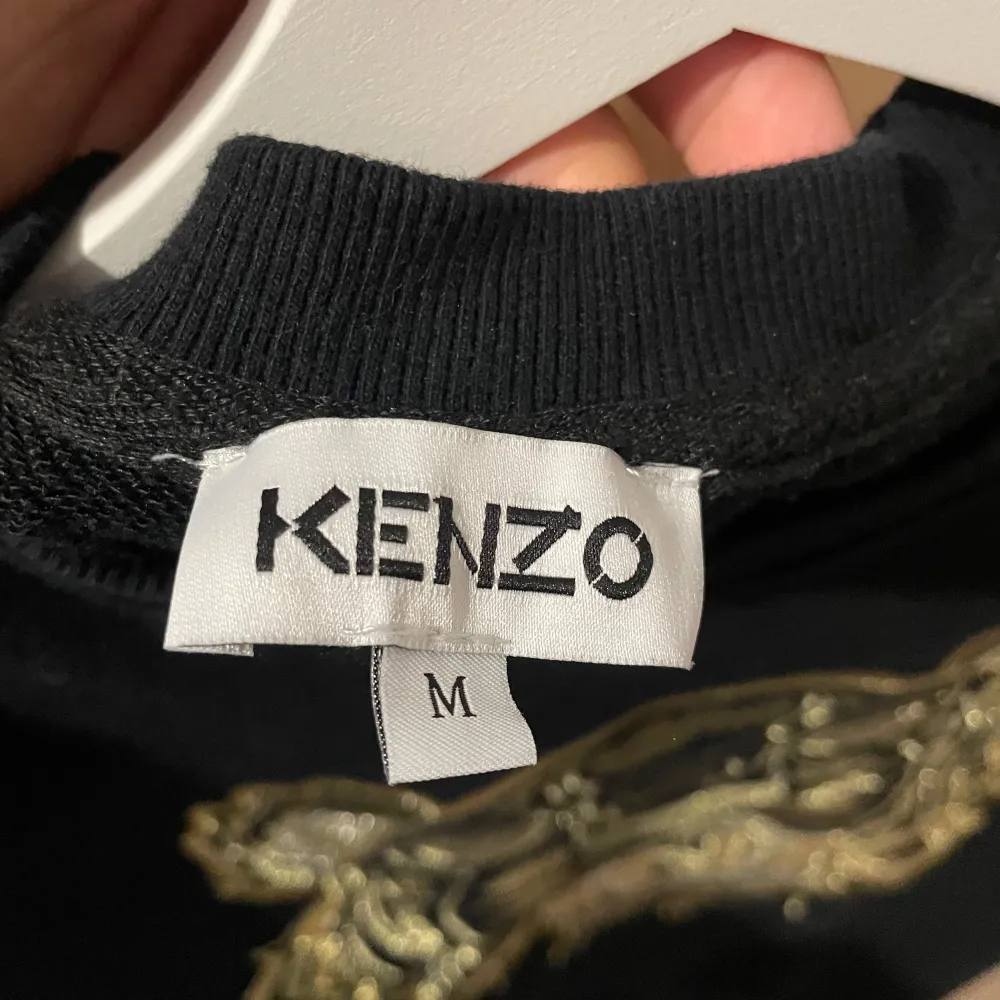 Säljer en svart sweatshirt från Kenzo med ett stort broderat tigerhuvud i guld på framsidan. Tröjan har långa ärmar och en rund halsringning. Perfekt för en stilren och trendig look.. Neuletakit & Villapaidat.